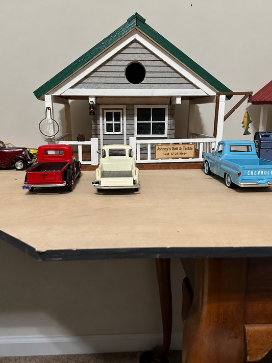 A miniature dollhouse-style birdhouse with a porch and several toy trucks displayed on top, photographed indoors against a plain wall.