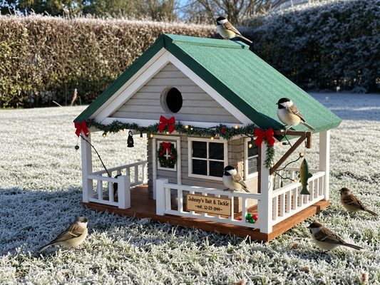 A decorative garden birdhouse shaped like a small house with a green roof, white porch, holiday garland, red bows, and a sign reading 'Johnny's Bait & Tackle Shop- and established 12-23-1944. Several birds are perched on and around the birdhouse in a frosty outdoor garden setting.