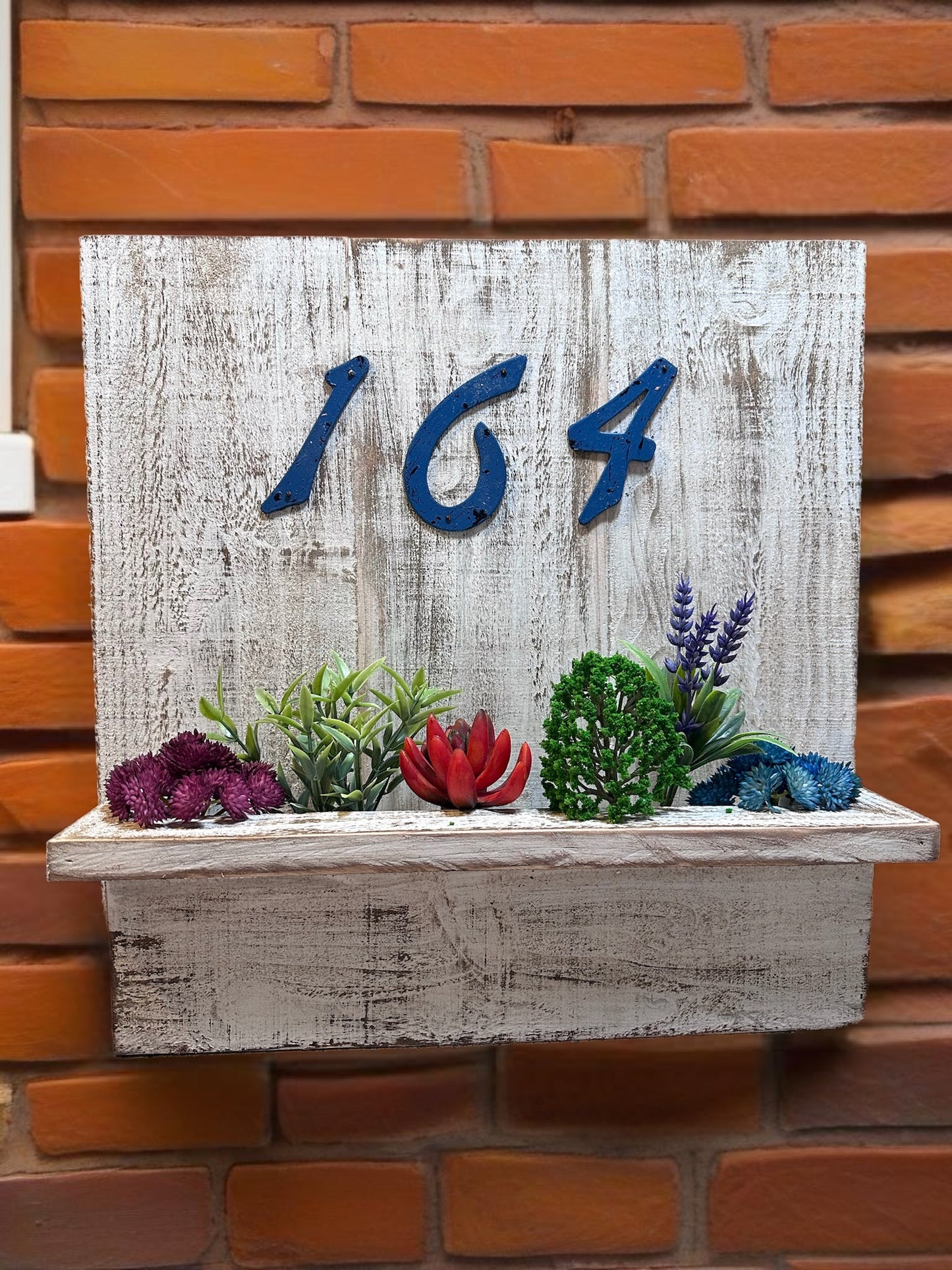House number planter box stained white displaying "621" in blue numbers, containing vibrant flowers and succulents, mounted on the siding of a house near the front door in bright sunlight.