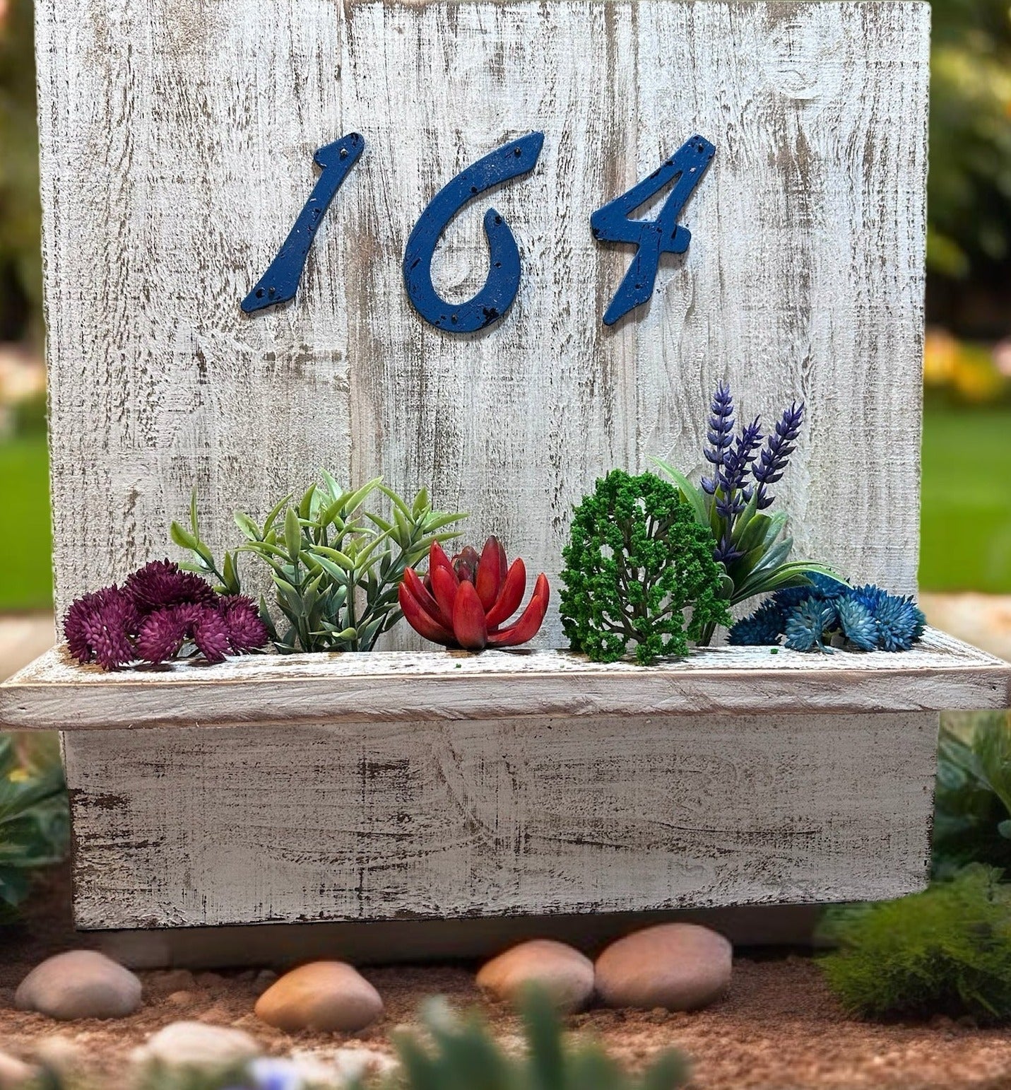 House number planter box stained white displaying "621" in blue numbers, containing vibrant flowers and succulents, mounted on the siding of a house near the front door in bright sunlight.