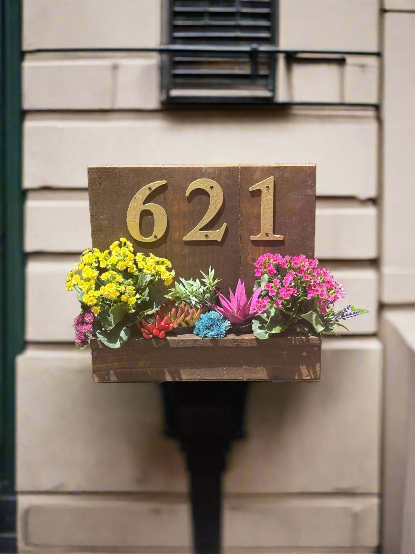 House number planter box displaying "621" in golden numbers, containing vibrant flowers and succulents, mounted on the siding of a house near the front door in bright sunlight.