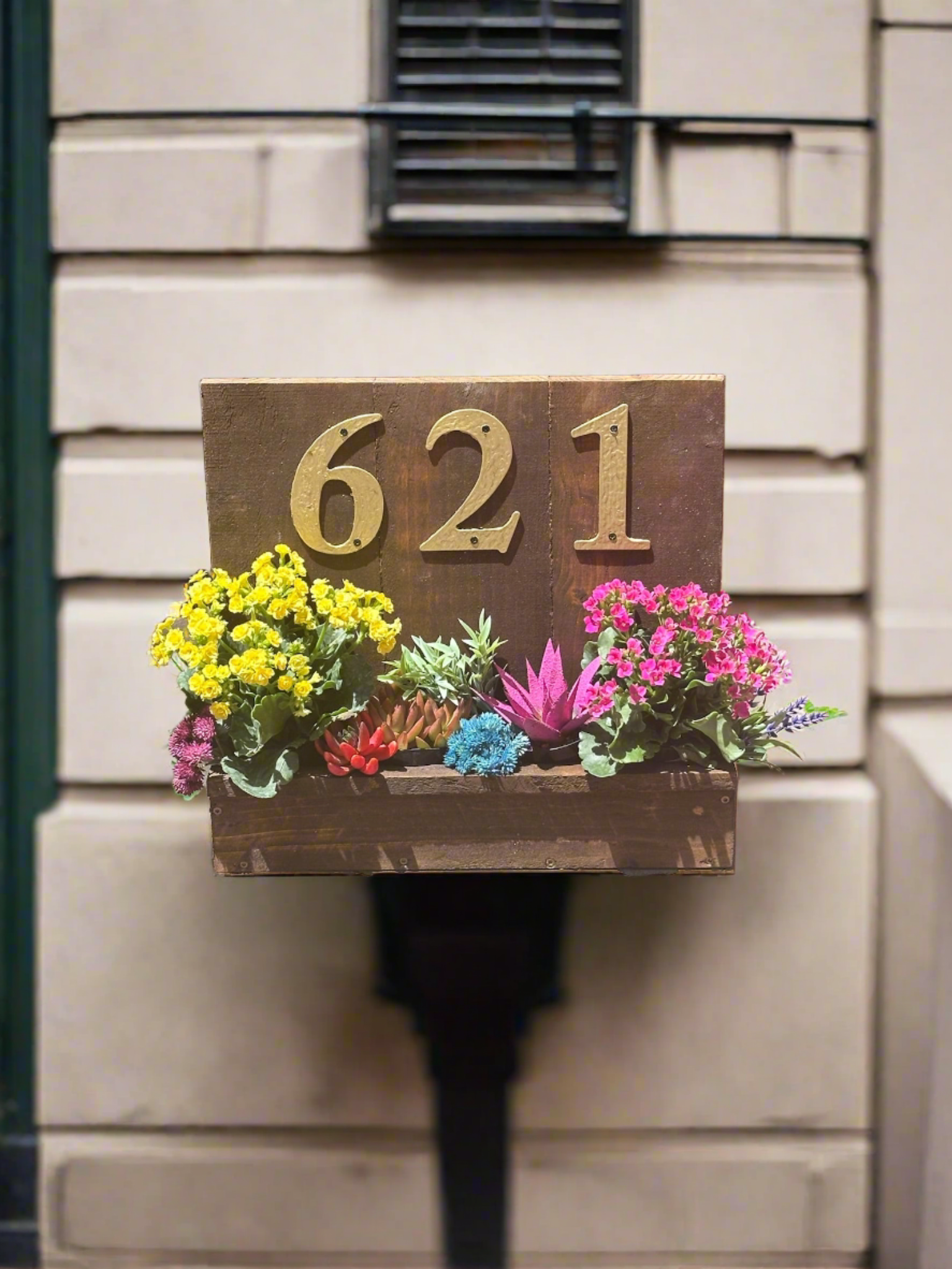 House number planter box displaying "621" in golden numbers, containing vibrant flowers and succulents, mounted on the siding of a house near the front door in bright sunlight.