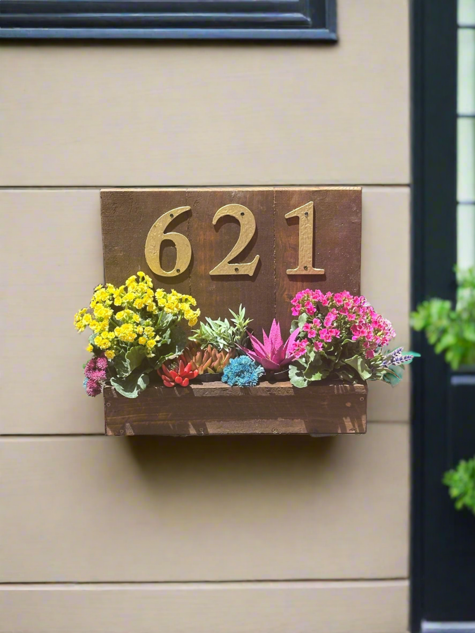 House number planter box displaying "621" in golden numbers, containing vibrant flowers and succulents, mounted on the siding of a house near the front door in bright sunlight.