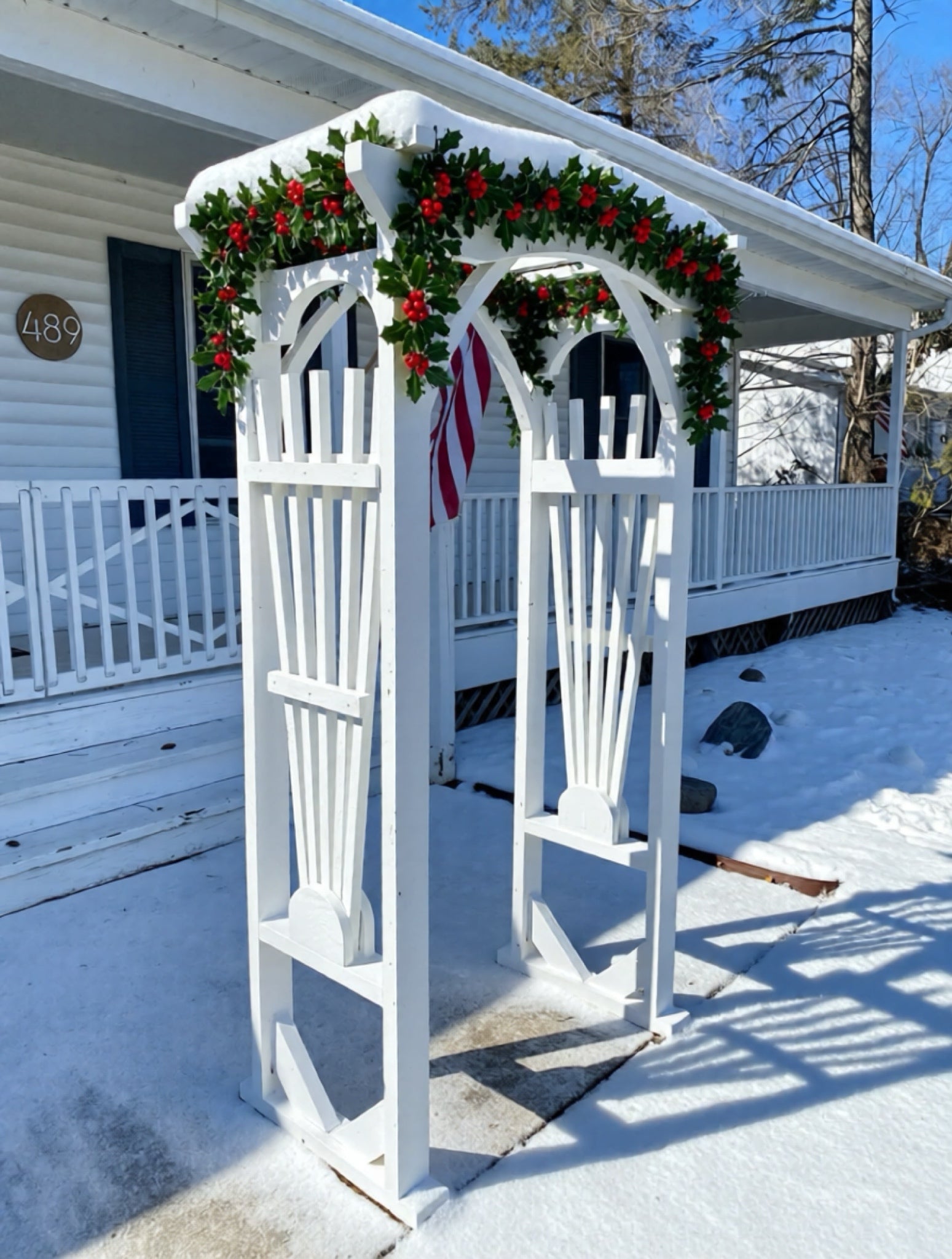 White decorative arch with Christmas garland and red berries on a snowy porch.