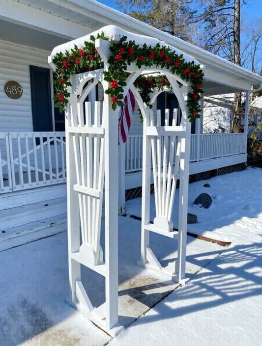 White decorative arch with Christmas garland and red berries on a snowy porch.