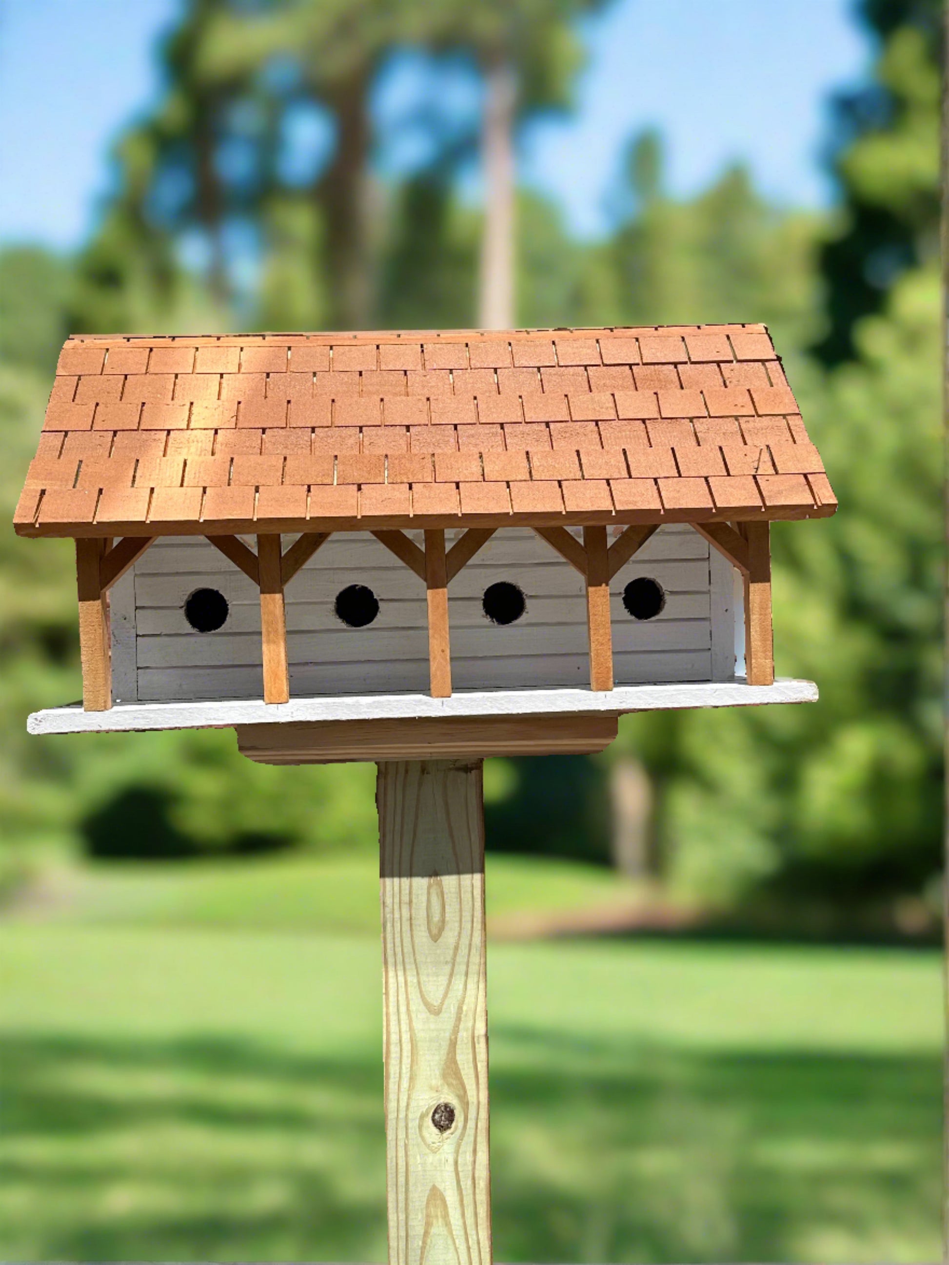 Purple Martin birdhouse with a shingled roof, mounted on a post in a sunny garden, surrounded by trees.