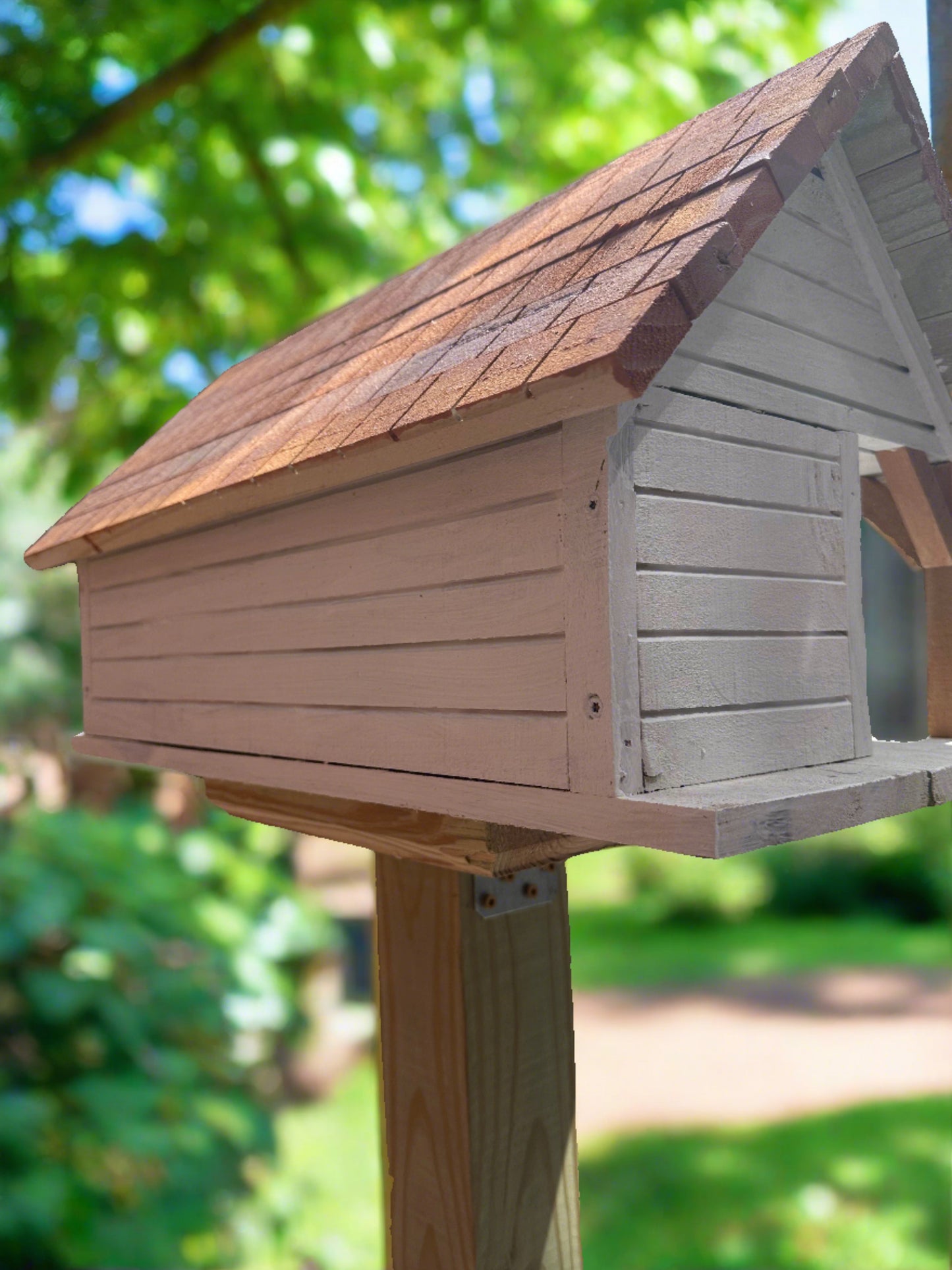 Purple Martin birdhouse with a shingled roof, mounted on a post in a sunny garden, surrounded by trees.