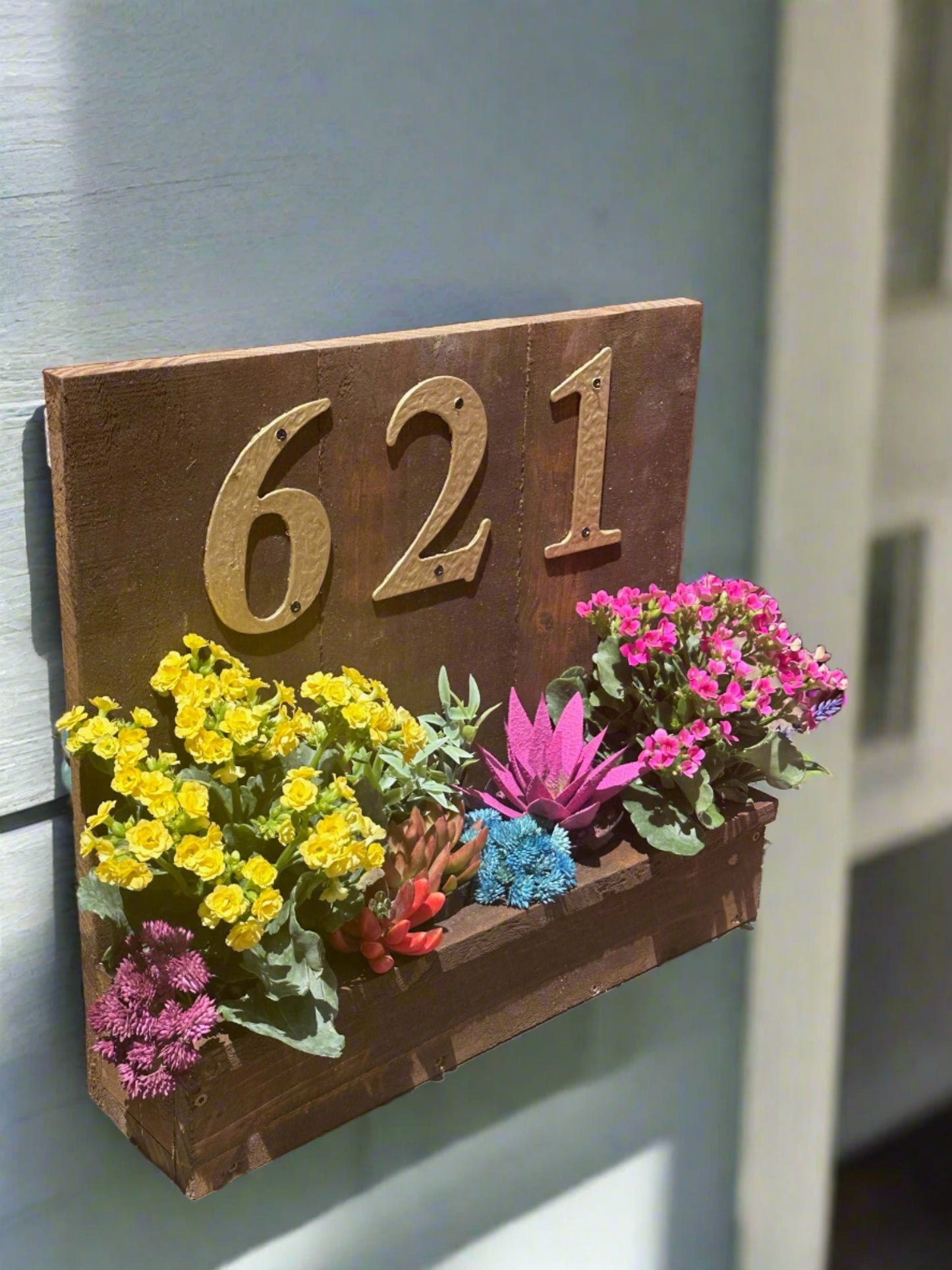 House number planter box displaying "621" in golden numbers, containing vibrant flowers and succulents, mounted on the siding of a house near the front door in bright sunlight.