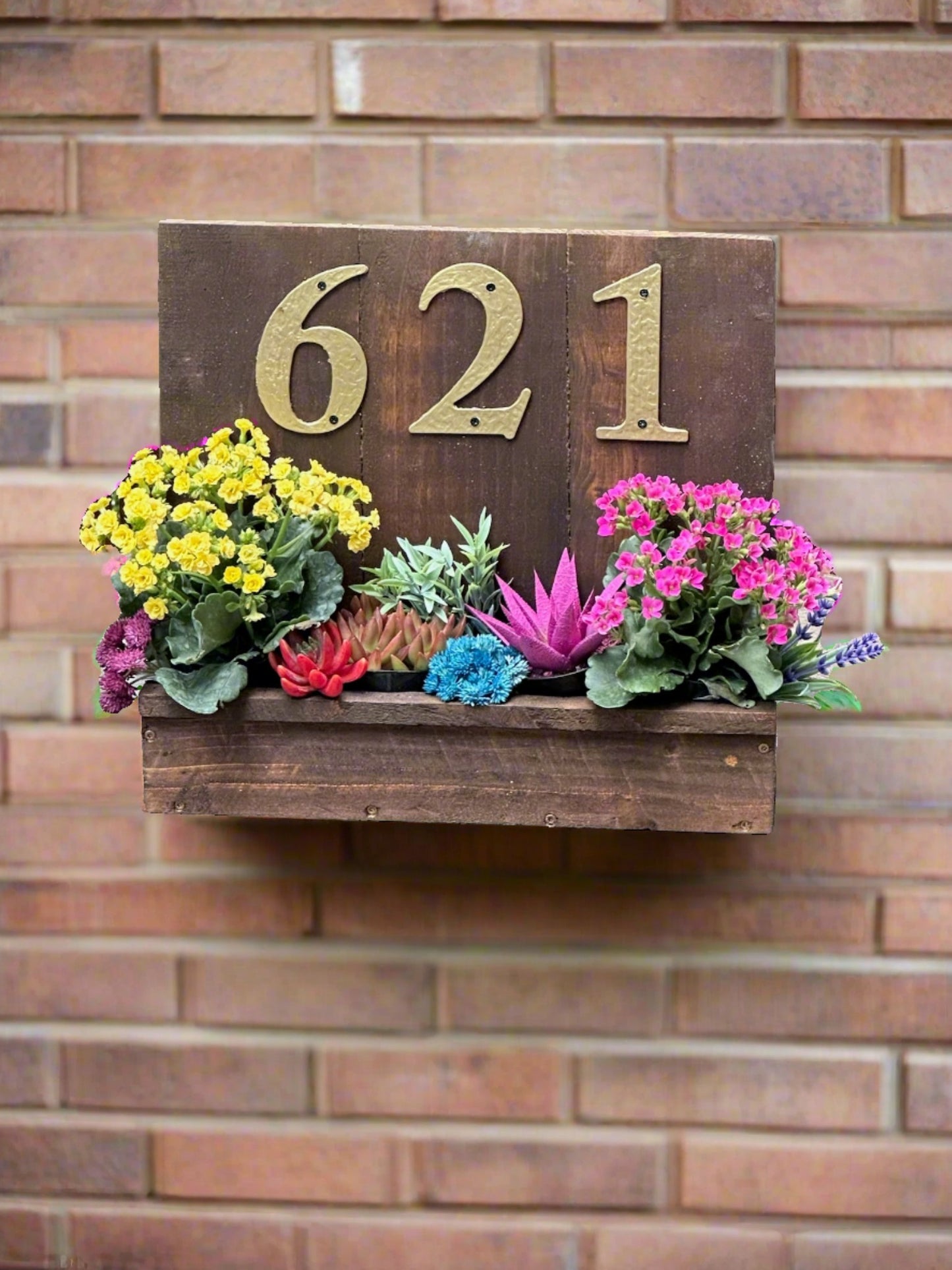 House number sign with golden numbers "621" hanging on a house front, featuring a planter box filled with colorful flowers and succulents, set against a brick wall background.