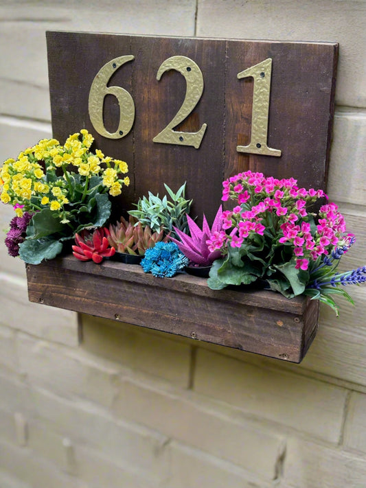 House number sign hanging on the siding next to the front door of a porch, featuring golden numbers "621" with a wooden planter box filled with colorful flowers and succulents, in bright sunlight.