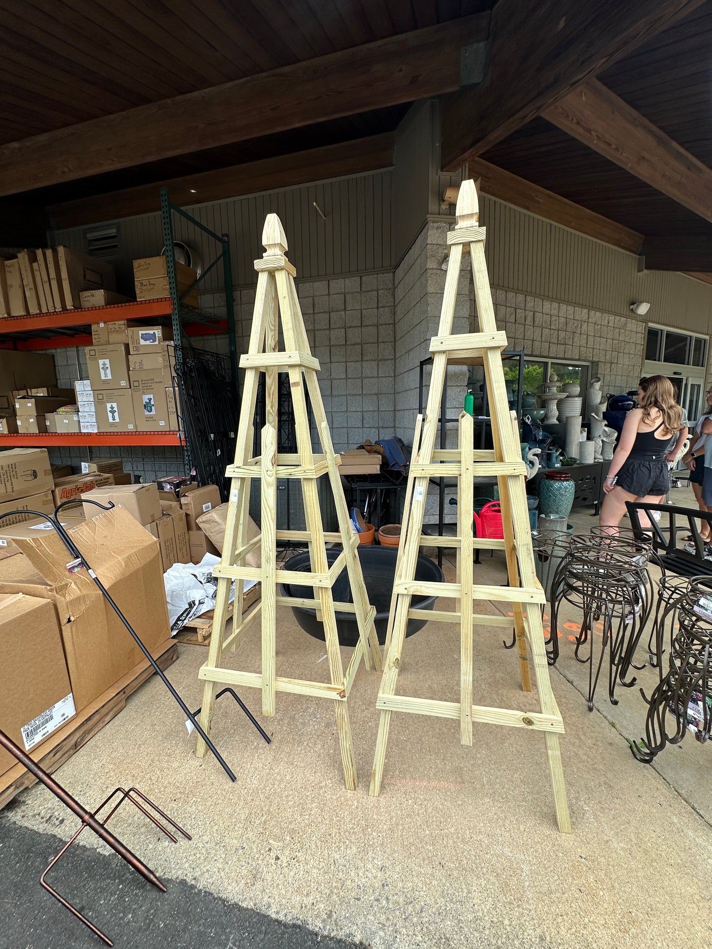 Two wooden obelisk garden trellises standing upright inside a storage area, with various items in the background.